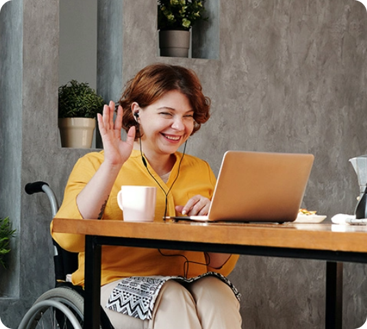 Zoom-out Picture of Woman in Yellow Crew Neck T-shirt Sitting on Black Wheelchair in front of Desk Waving to Laptop