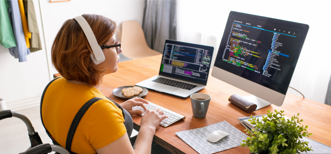 A person in a wheelchair wearing headphones writes code on a laptop and external monitor.