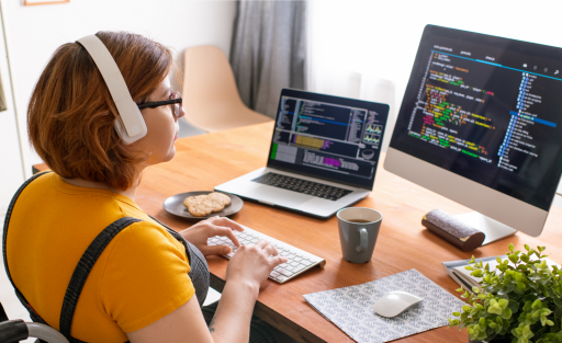A person in a wheelchair wearing headphones writes code on a laptop and external monitor.